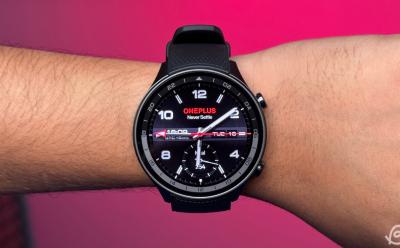 OnePlus 2R on a person's wrist with an analog watch face in front of a bright pink background