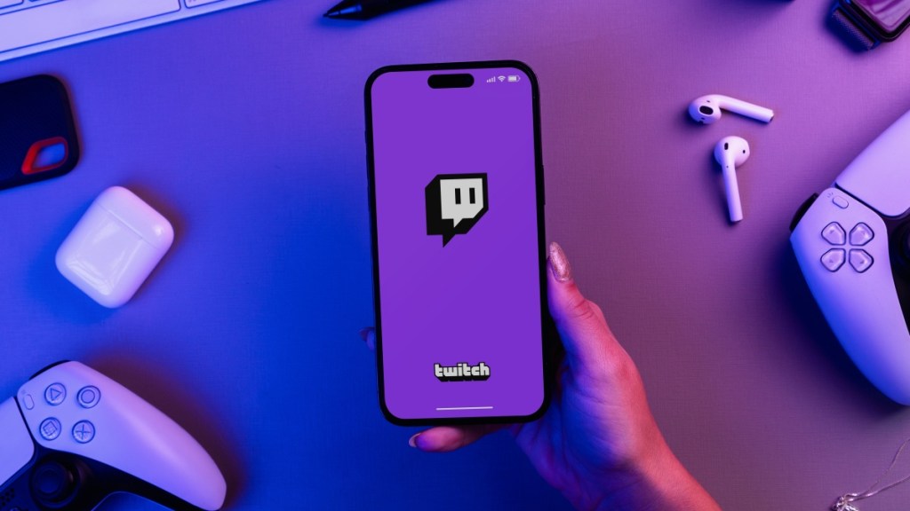 Girl holding an iPhone 14 Pro smartphone with Twitch app with PS5 video game controllers.