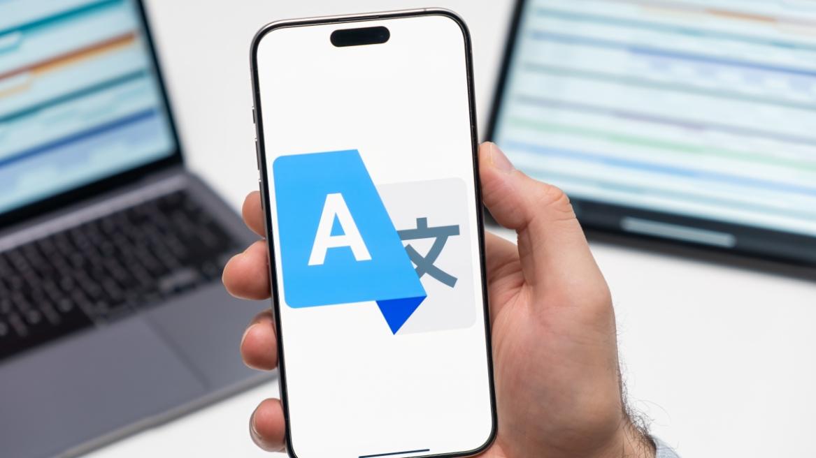 Google Translate logo on the screen of smartphone in mans hand and laptop on the background